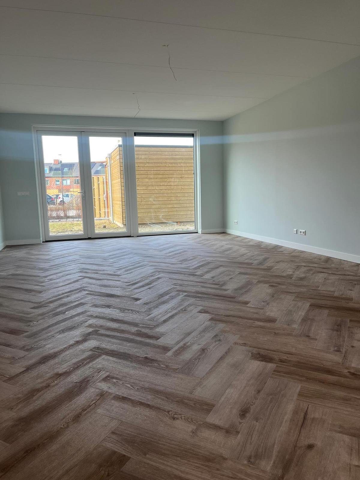 Ruime, lichte woonkamer met openslaande tuindeuren en volledig gelegde visgraat PVC-vloer, iets ander perspectief.