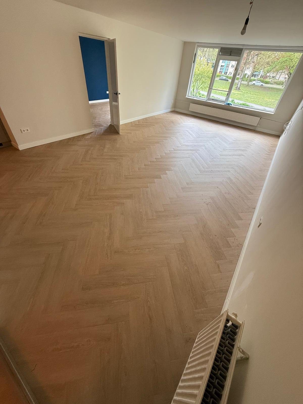 Ruime woonkamer met groot hoekraam en uitzicht op een park, volledig gelegde lichte visgraat PVC-vloer.