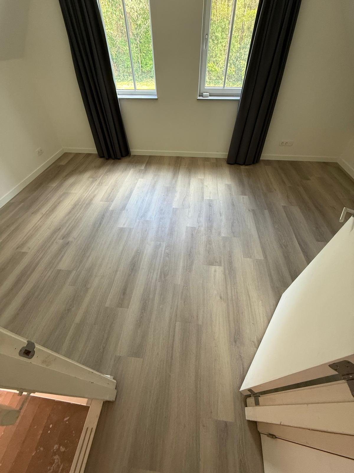 Slaapkamer met hoge ramen en donkere gordijnen, volledig gelegde rechte plankenvloer, trap deels zichtbaar.